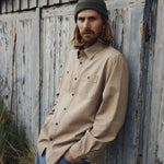 Man wearing a beige shirt and blue jeans leaning against a rustic wooden wall.