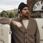 Man wearing a brown cardigan and black beanie leaning against a vintage vehicle with trees and signs in the background.