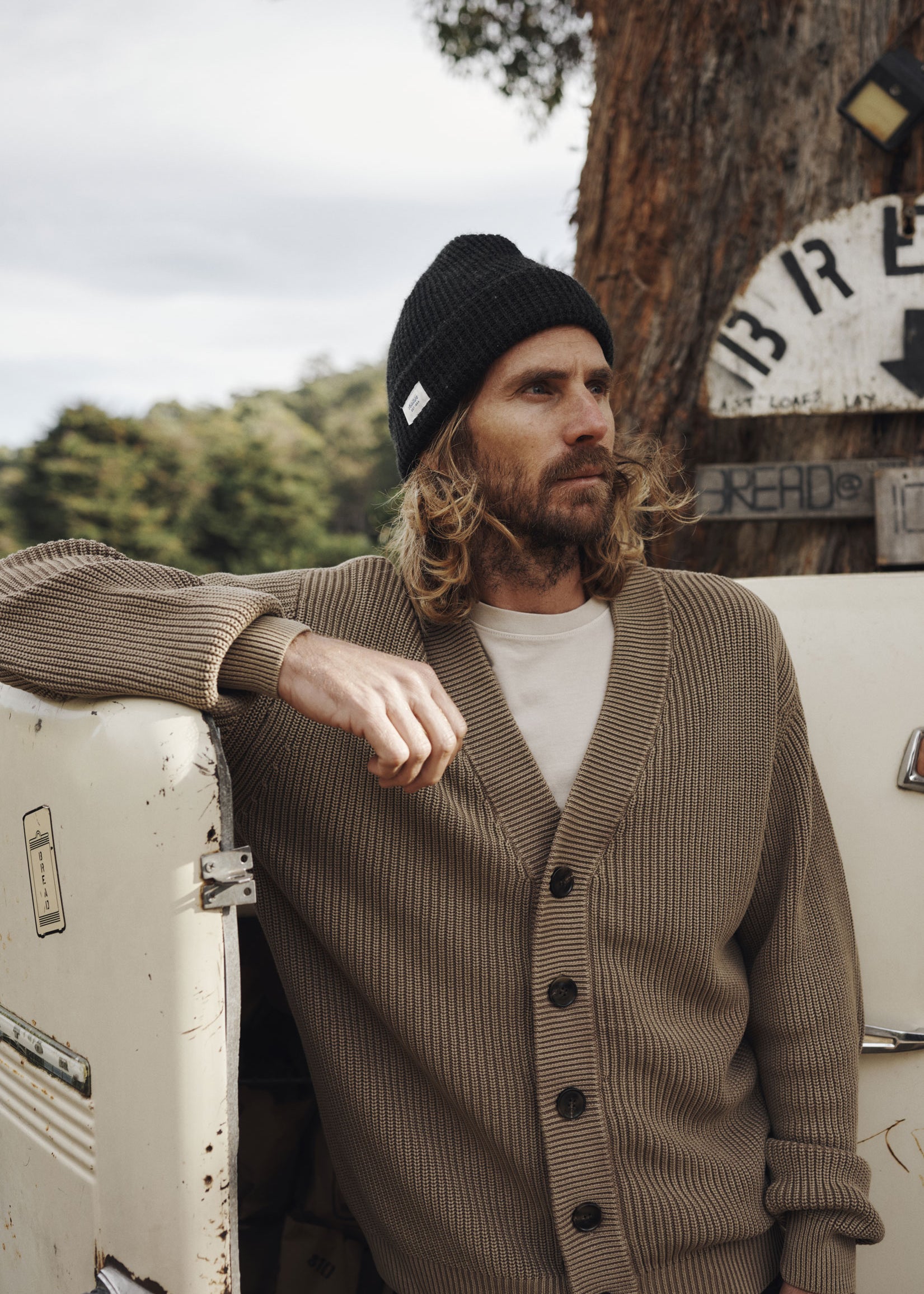 Man wearing a brown cardigan and black beanie leaning against a vintage vehicle with trees and signs in the background.