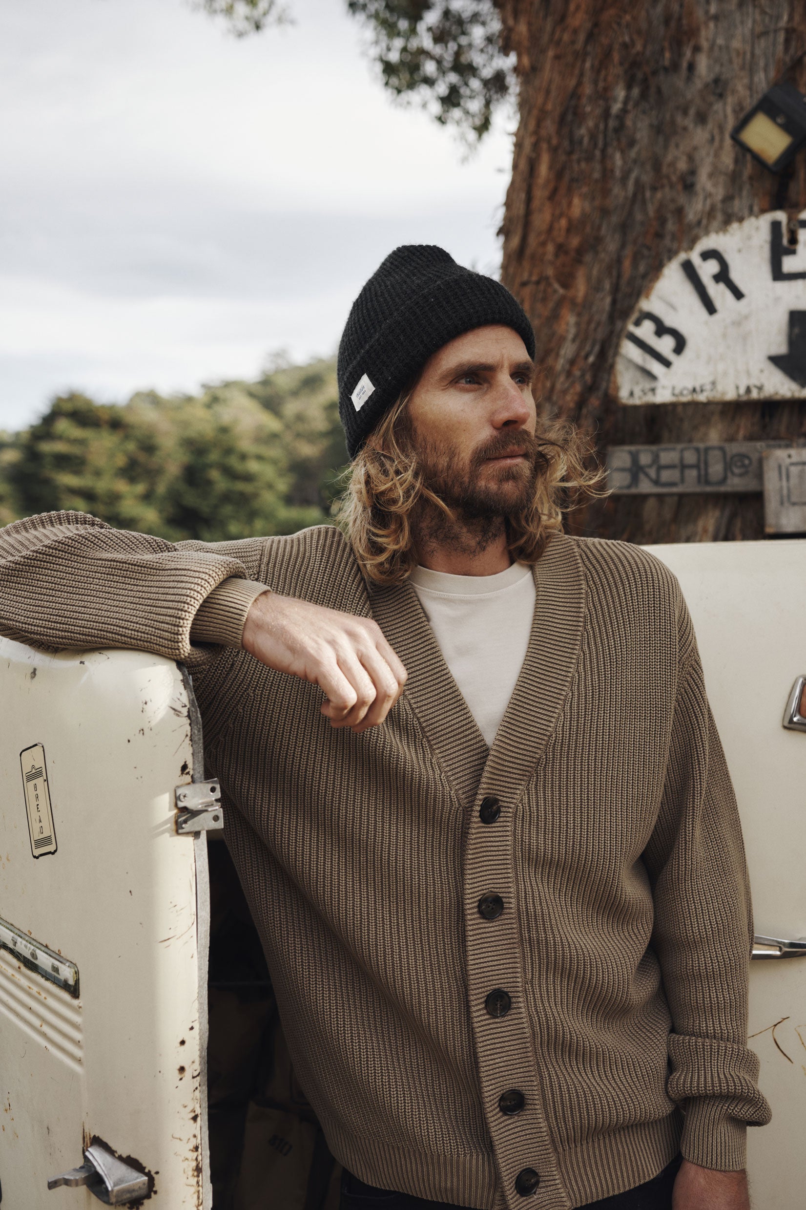 Man wearing a brown cardigan and black beanie leaning against a vintage vehicle with trees and signs in the background.