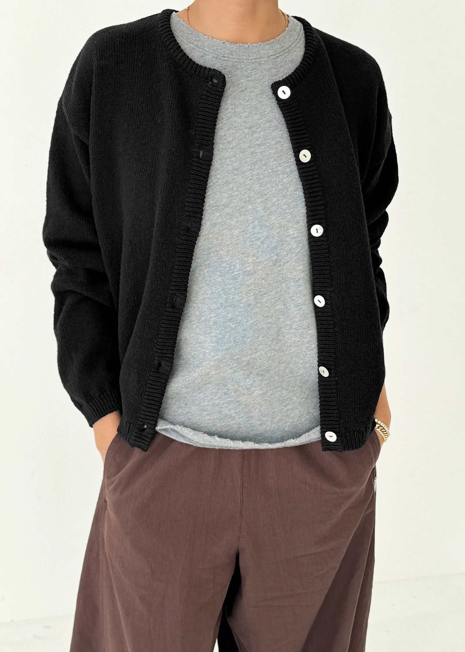 Person wearing a black cardigan over a gray shirt with brown pants on a white background