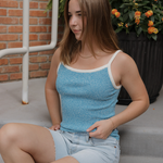 Woman sitting on a stairs wearing a blue tank top and denim shorts, with a potted plant in the background.