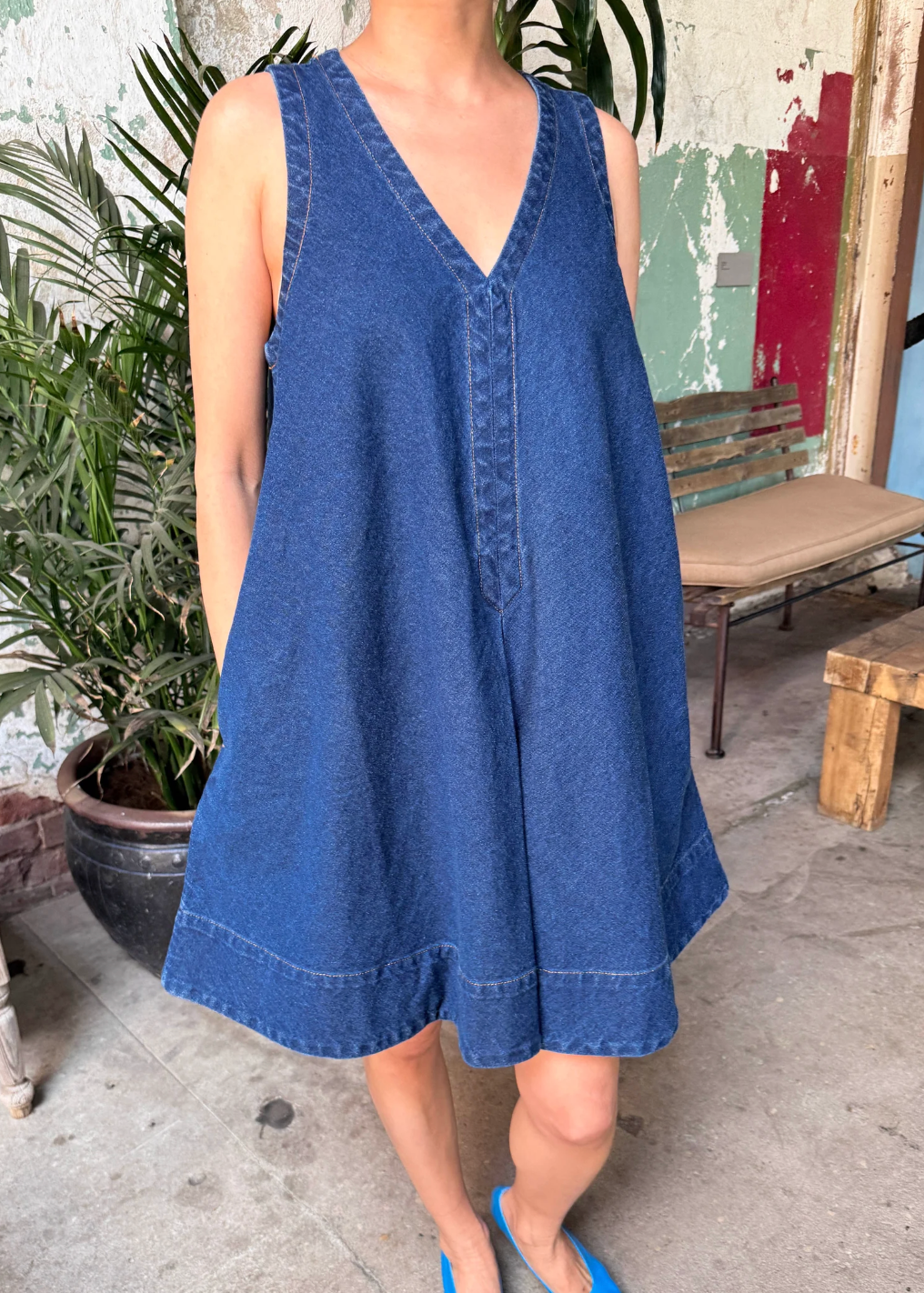 Person wearing a blue dress standing in an outdoor setting with plants and a bench.