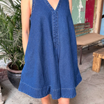 Person wearing a blue dress standing in an outdoor setting with plants and a bench.