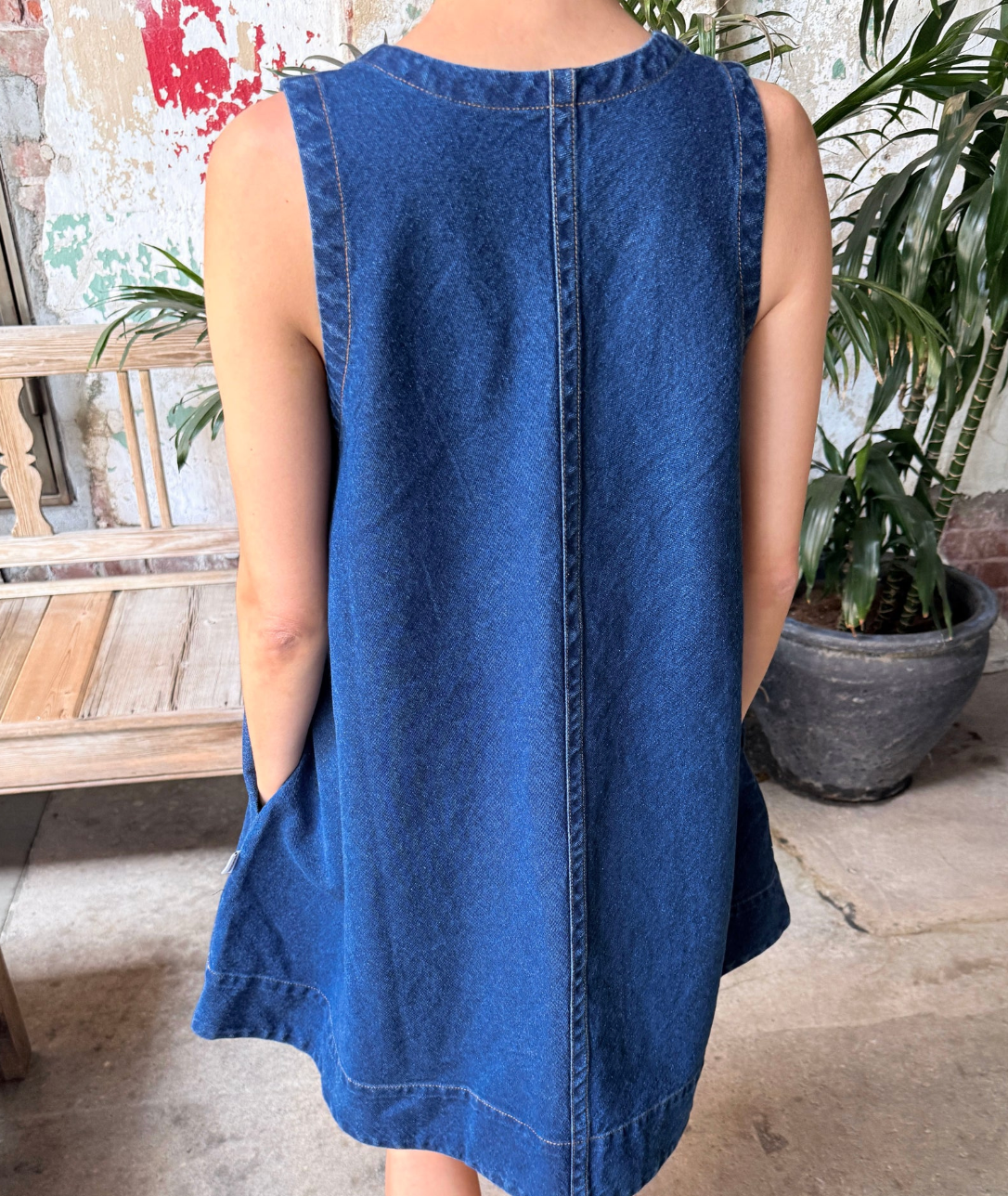 Person wearing a blue denim dress in an outdoor setting with plants and a wooden bench.