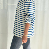 Person wearing a blue and white striped shirt with rolled-up sleeves, standing against a white curtain.