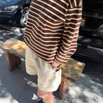 Person wearing a brown and white striped shirt and beige shorts standing on a sidewalk with cars in the background.