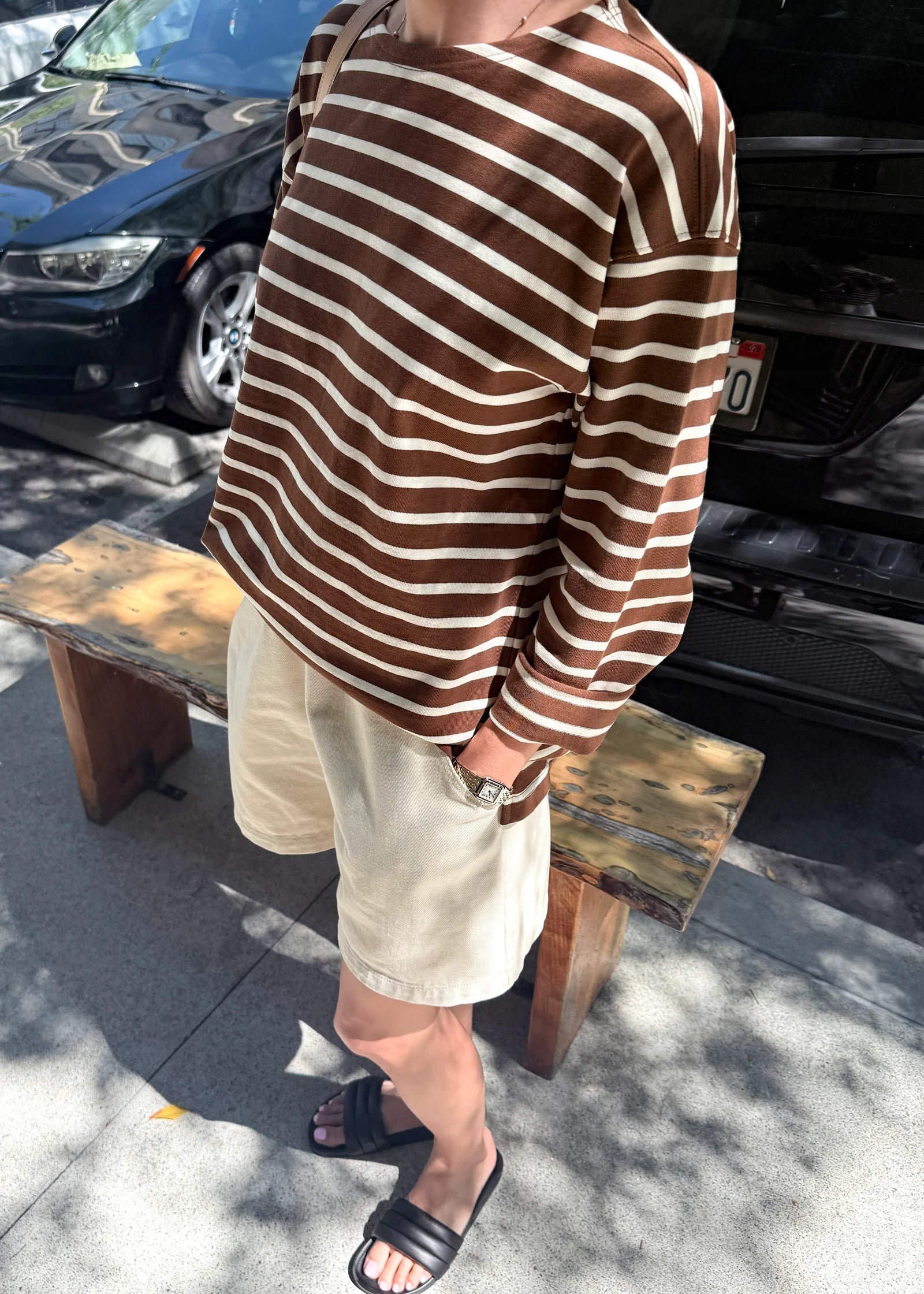 Person wearing a brown and white striped shirt and beige shorts standing on a sidewalk with cars in the background.
