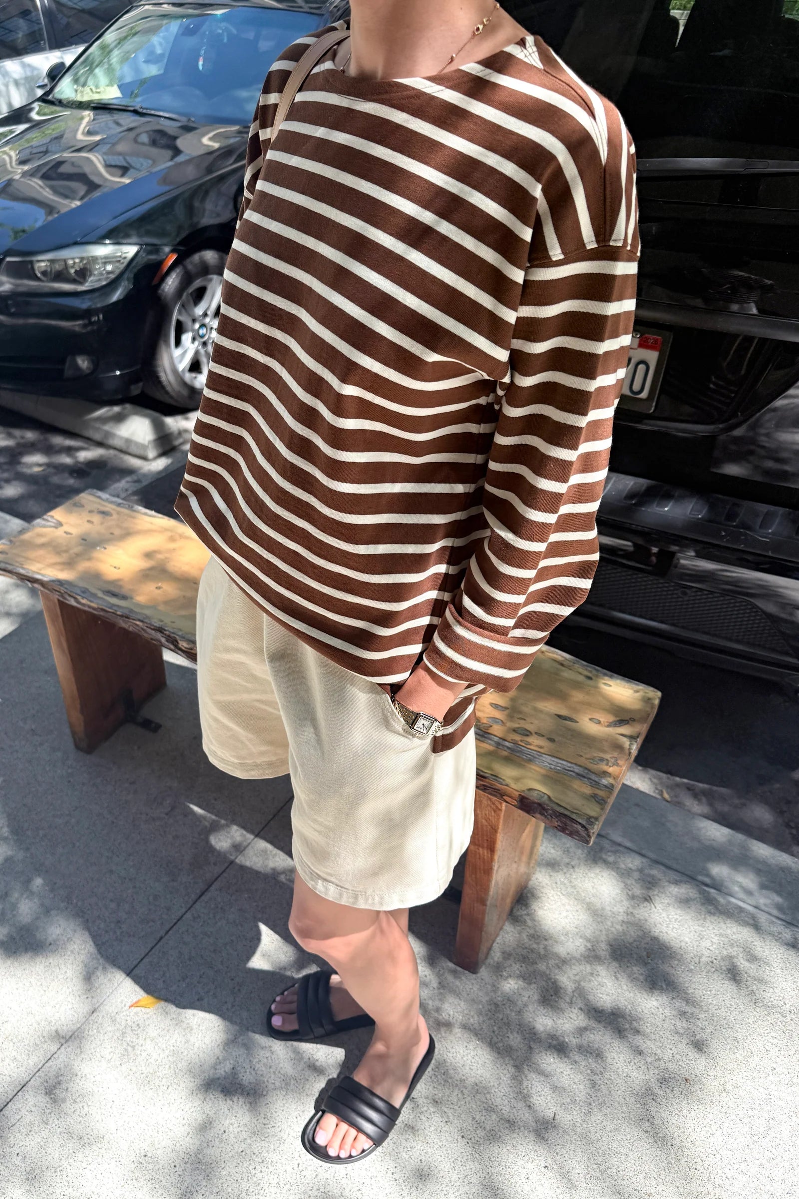 Person wearing a brown and white striped shirt and beige shorts standing on a sidewalk with cars in the background.
