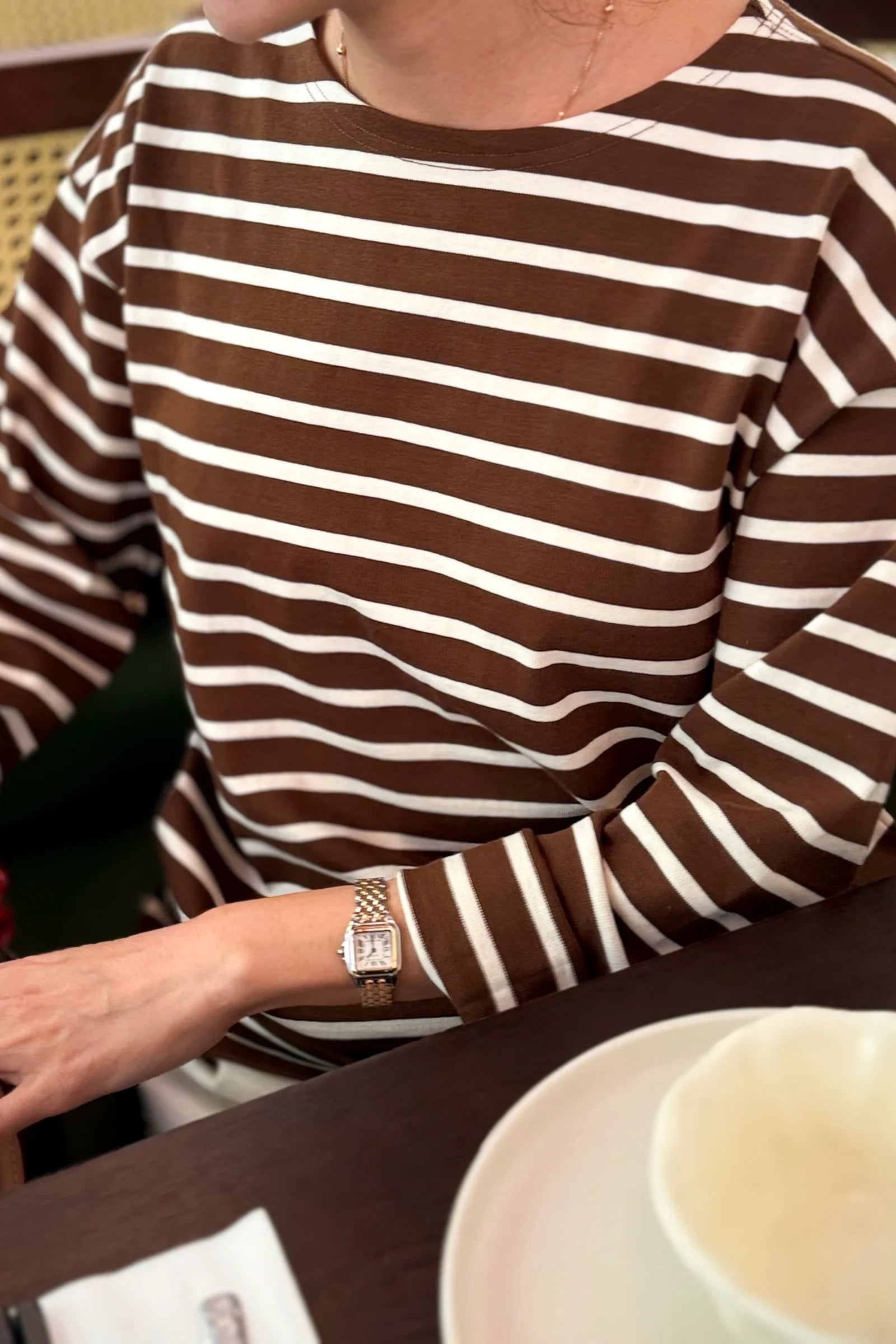 Person wearing a brown and white striped shirt with a watch, sitting at a table with a cup.