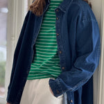 A person wearing a dark blue denim shirt over a green striped t-shirt paired with white pants.