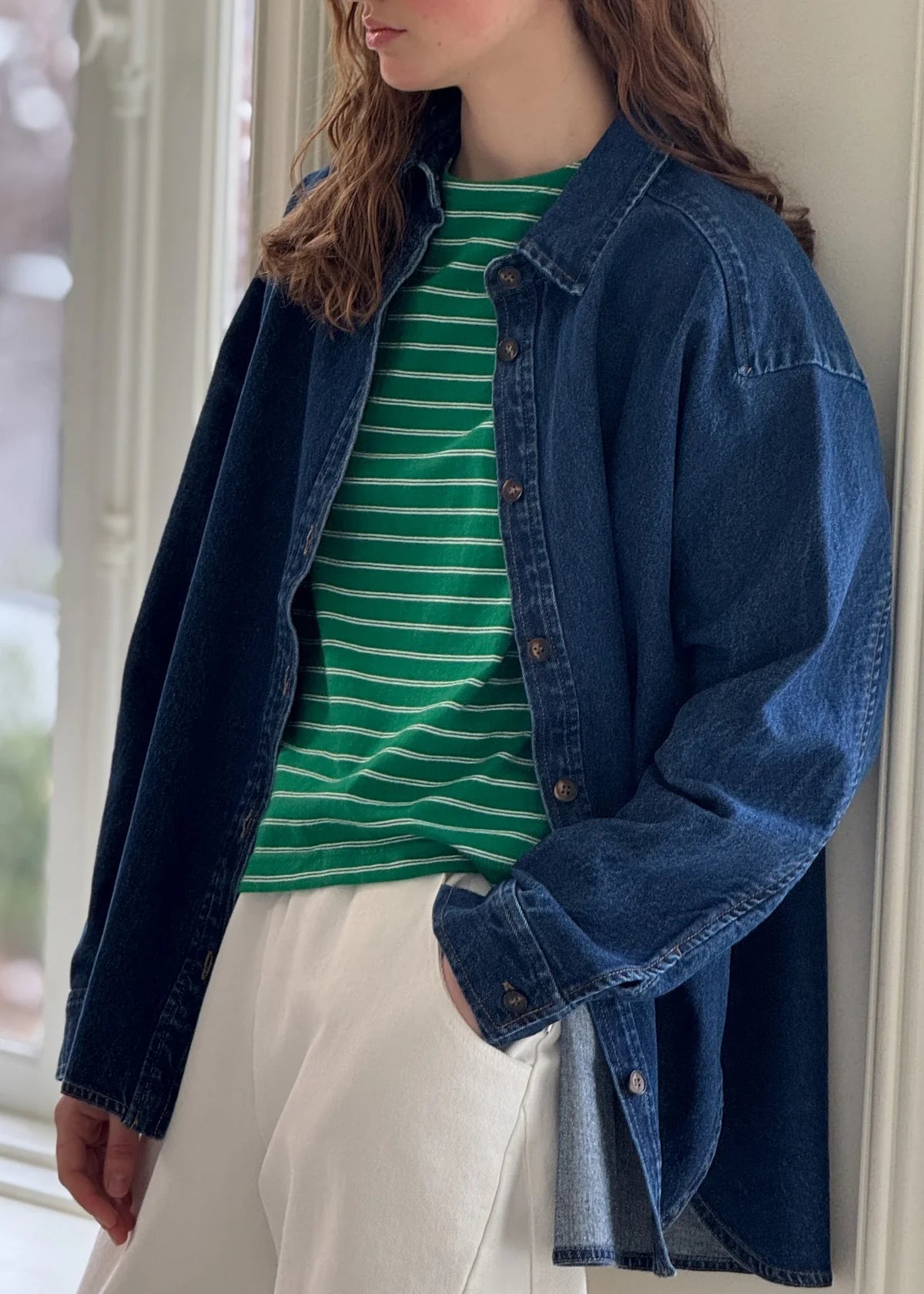 A person wearing a dark blue denim shirt over a green striped t-shirt paired with white pants.
