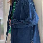 Woman wearing a green and white striped shirt with open dark denim shirt over it and white pants leaning against a white wall
