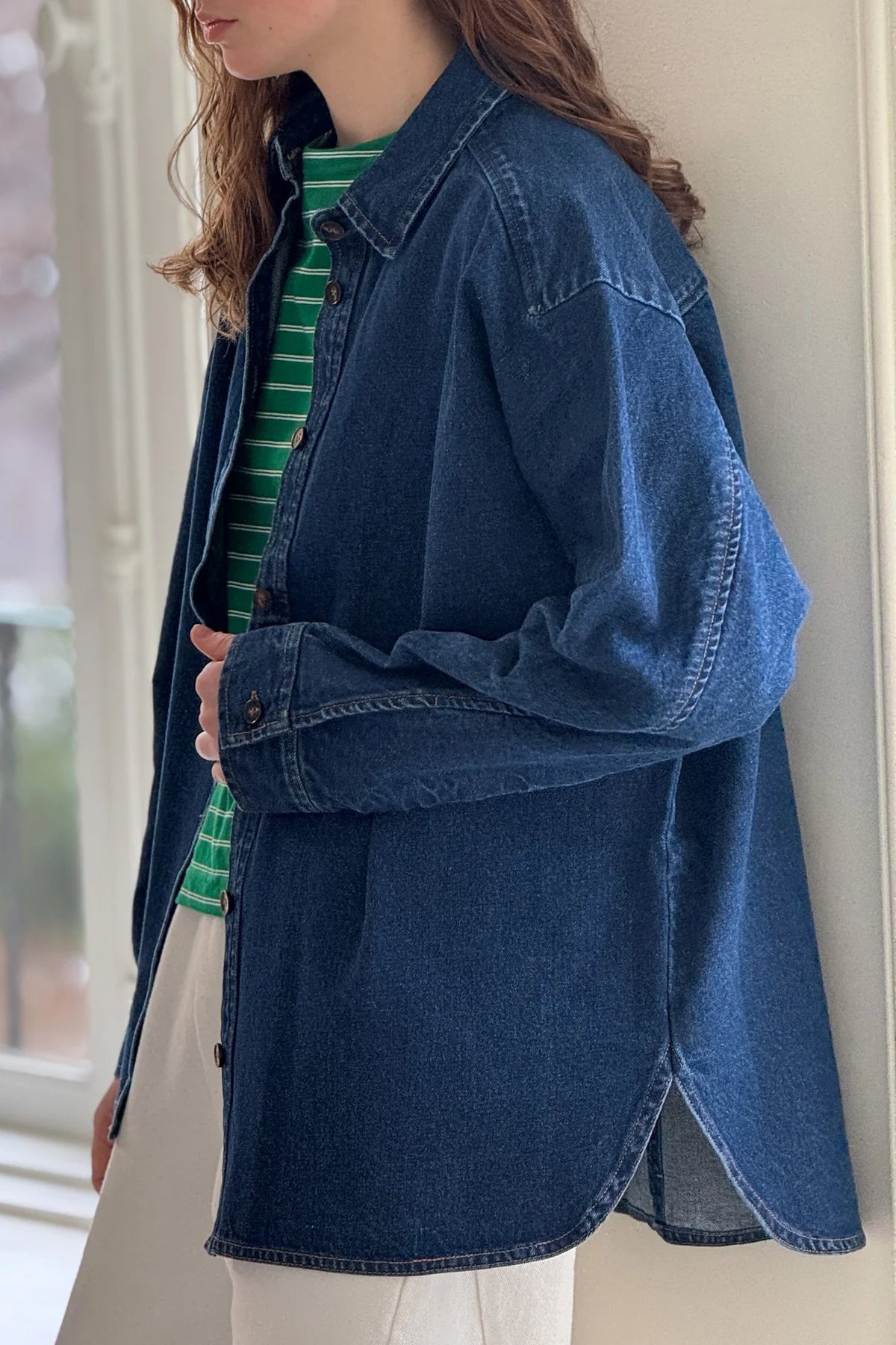 Woman wearing a green and white striped shirt with open dark denim shirt over it and white pants leaning against a white wall