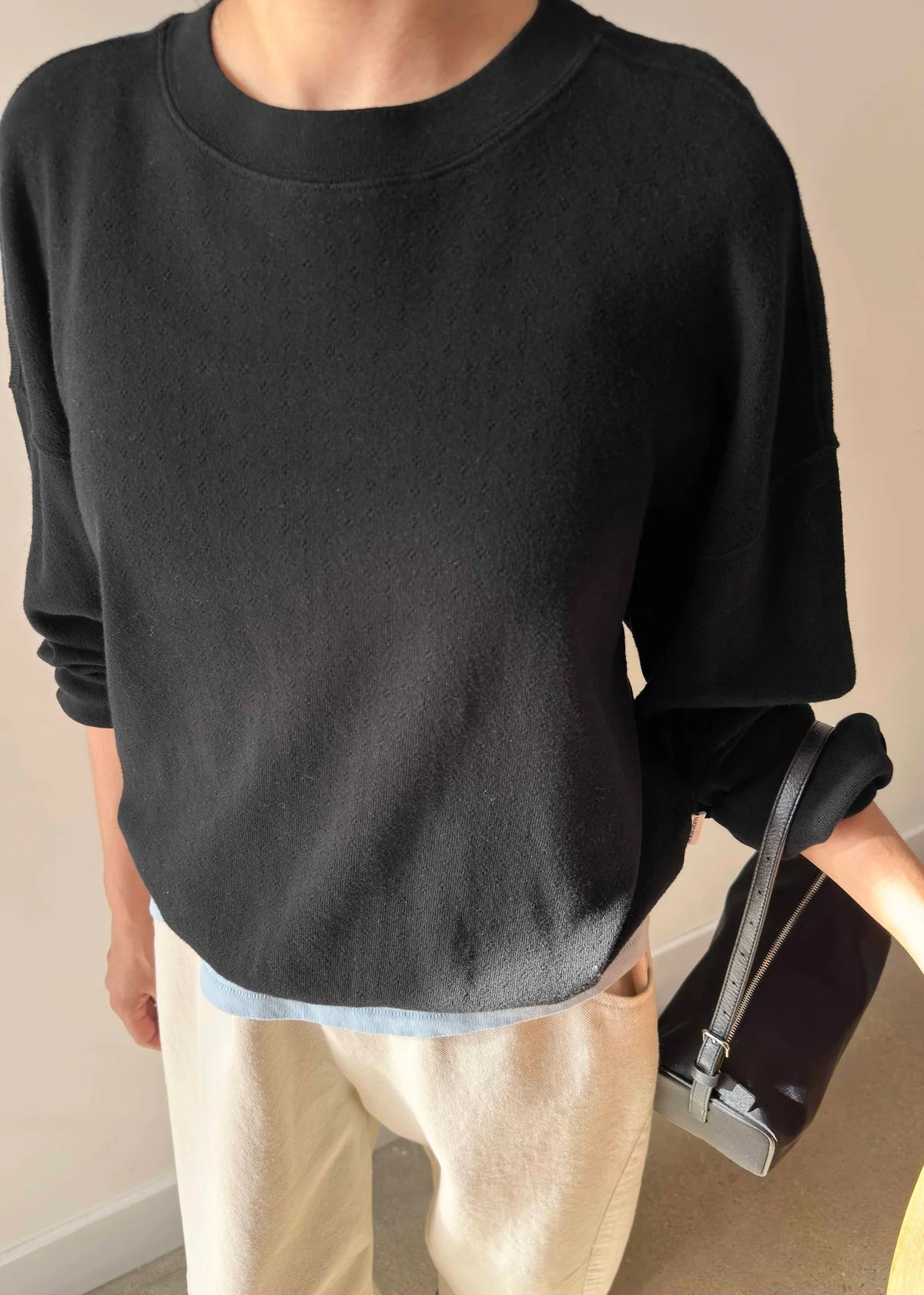 Person wearing a black sweater and beige pants holding a black handbag indoors.