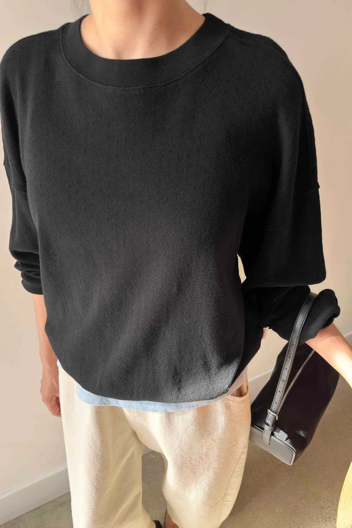Person wearing a black sweater and beige pants holding a black handbag indoors.