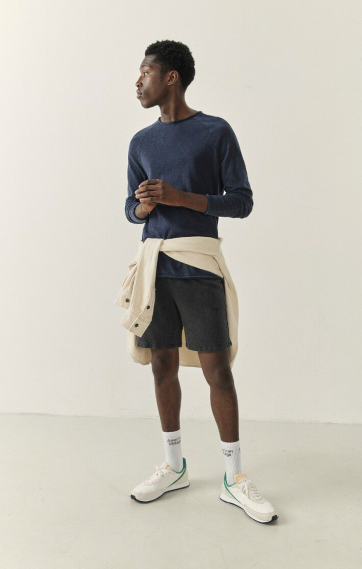 A full body image of a male model wearing the Sonoma Long Sleeve in vintage navy styled with black shorts and a cream shirt tied around his waist