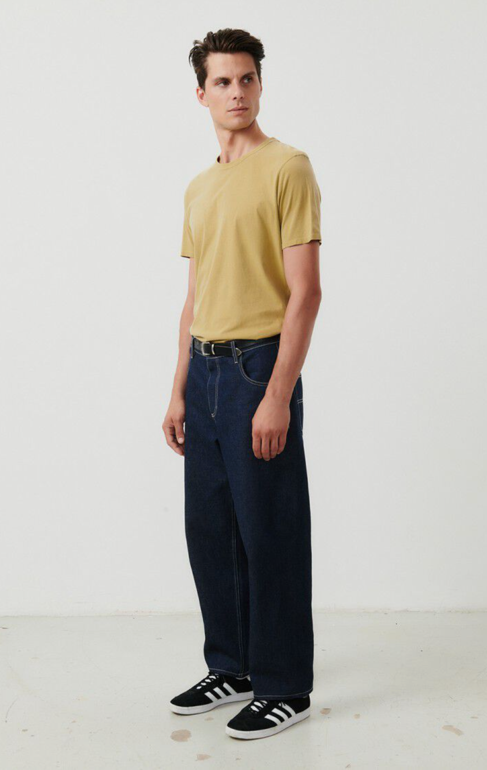 A full body front facing image of a male model standing at an angle while wearing the Devon t-shirt in vintage safari tucked into dark denim