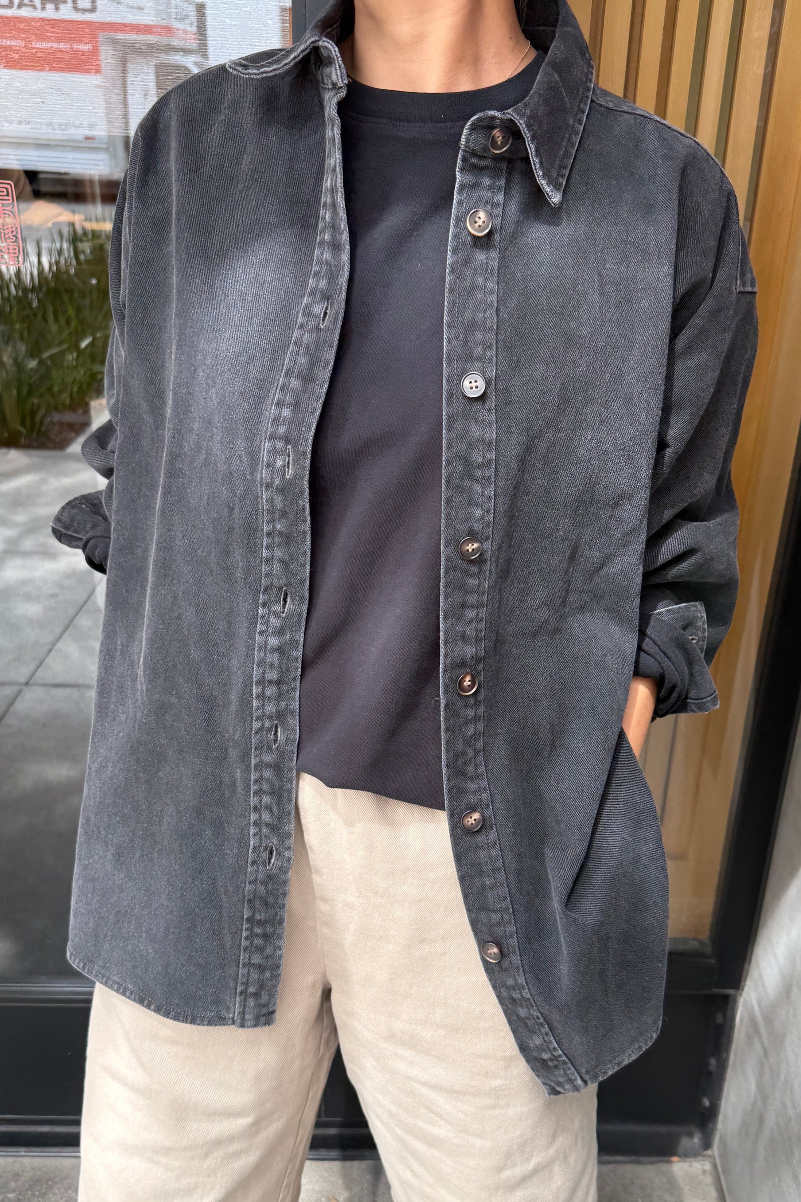 Person wearing a dark denim shirt over a black t-shirt with beige pants, standing indoors.