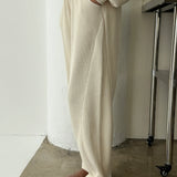 Person wearing a beige outfit with wide-leg pants and brown shoes in a minimal indoor setting.