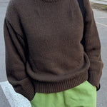 Person wearing a brown sweater and green pants on a street.