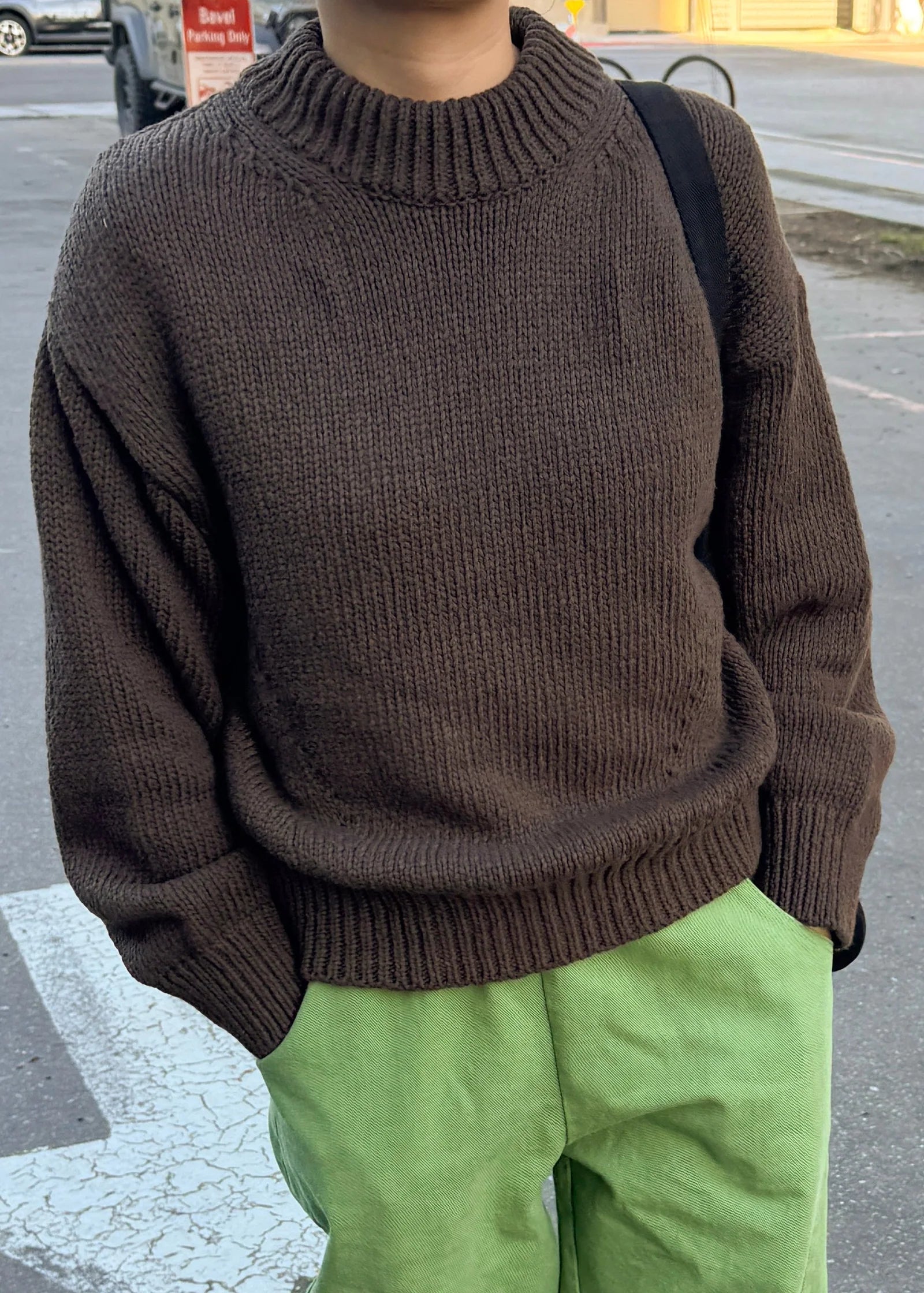 Person wearing a brown sweater and green pants on a street.