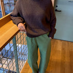 Person wearing a brown sweater and green pants standing indoors.