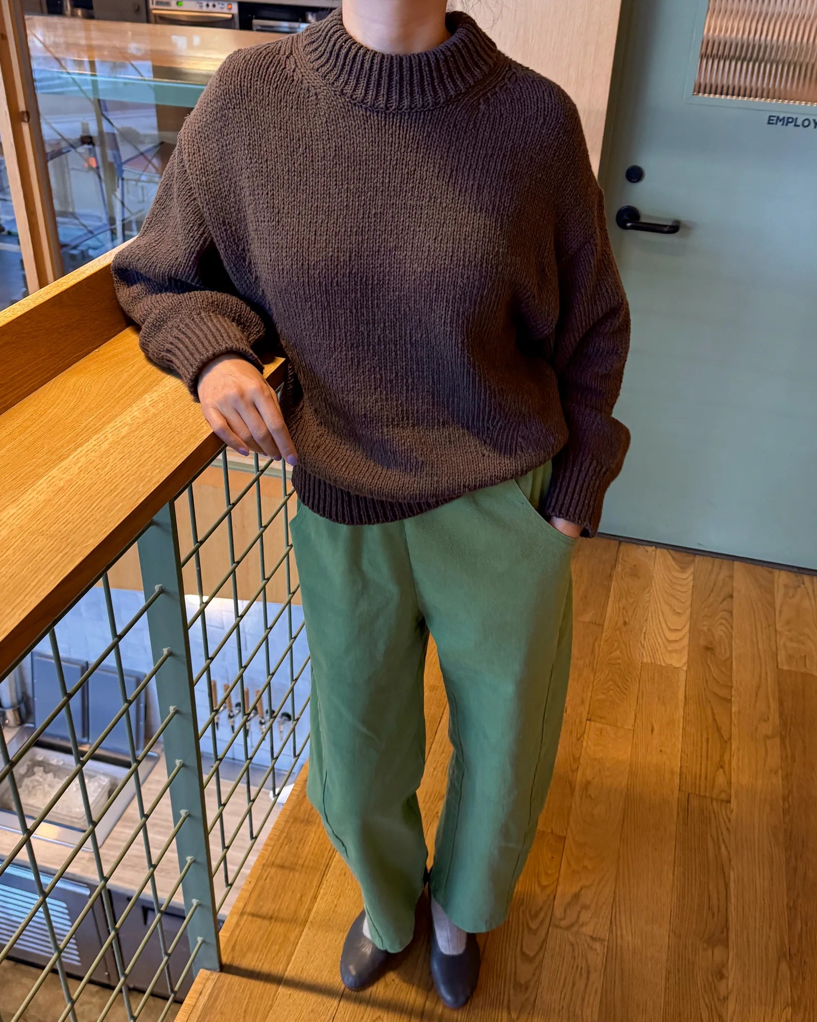 Person wearing a brown sweater and green pants standing indoors.