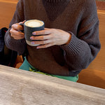 Person wearing a brown sweater holding a mug with a brick wall background