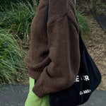 Person wearing a brown sweater and green pants, holding a black bag with white text.
