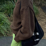 Person wearing a brown sweater and green pants, holding a black bag with white text.