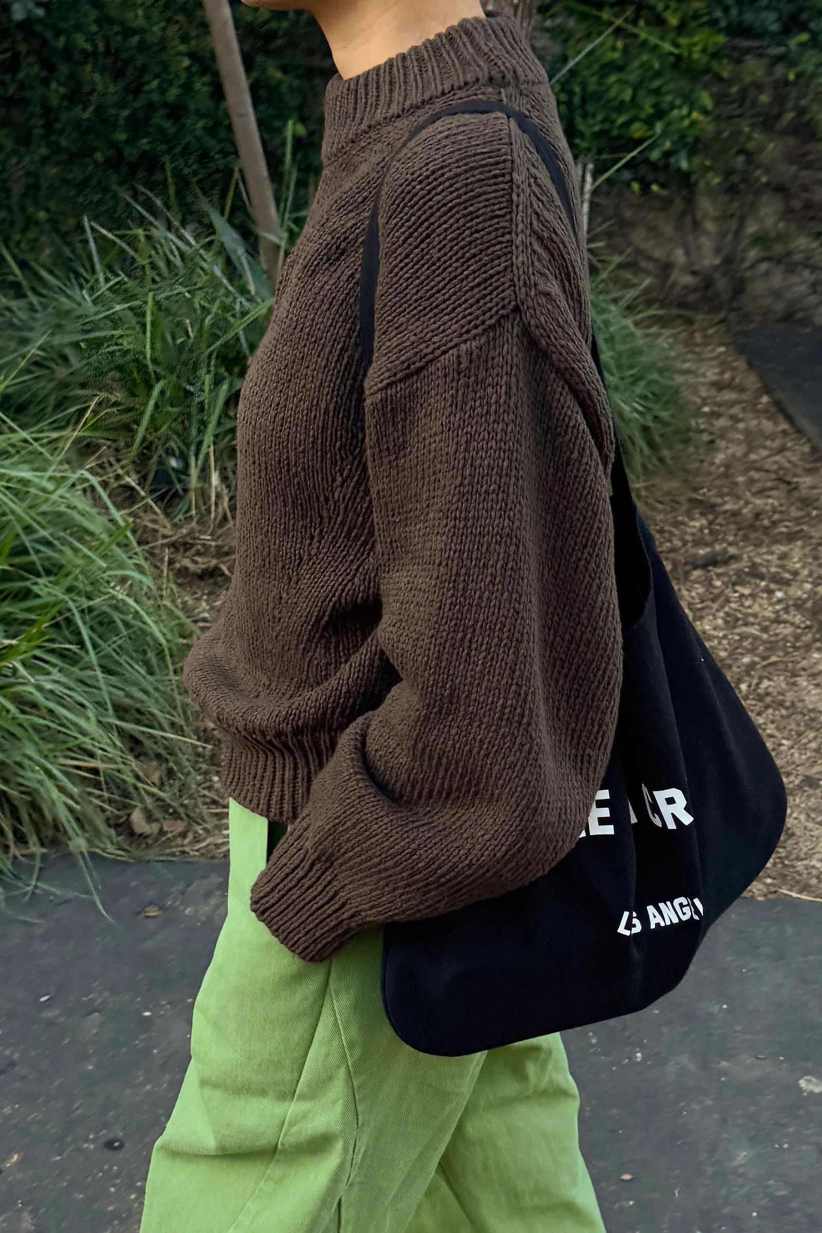 Person wearing a brown sweater and green pants, holding a black bag with white text.