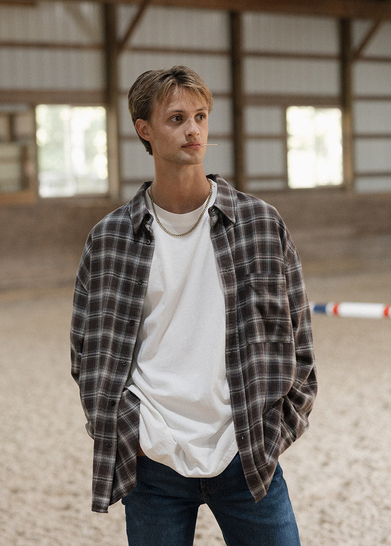 Man standing in an indoor equestrian arena wearing a plaid shirt over a white t-shirt.