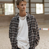 Man standing in an indoor equestrian arena wearing a plaid shirt over a white t-shirt.