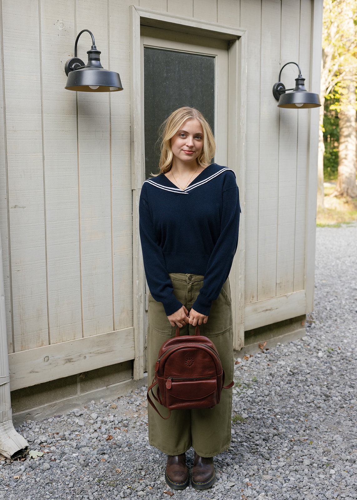 Woman standing outside a wooden building holding a brown backpack