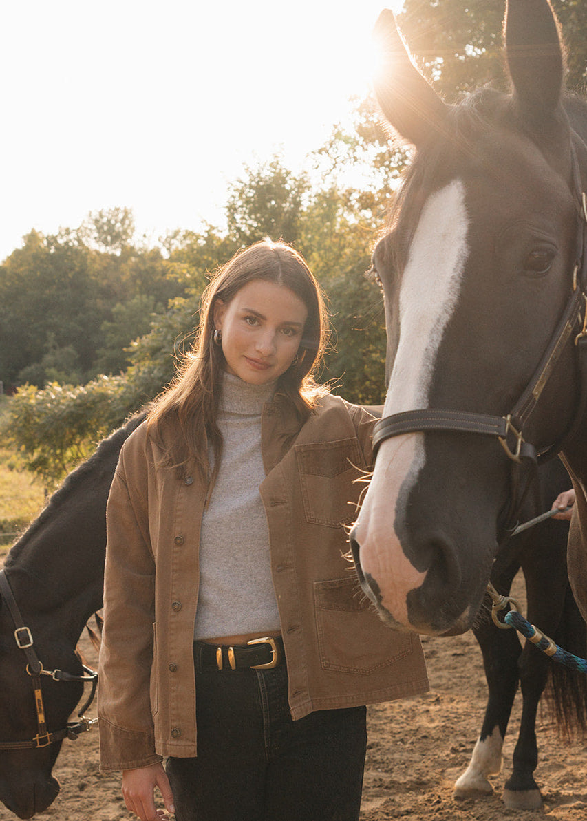 Woman standing with a horse in an outdoor setting with trees and a barn in the background