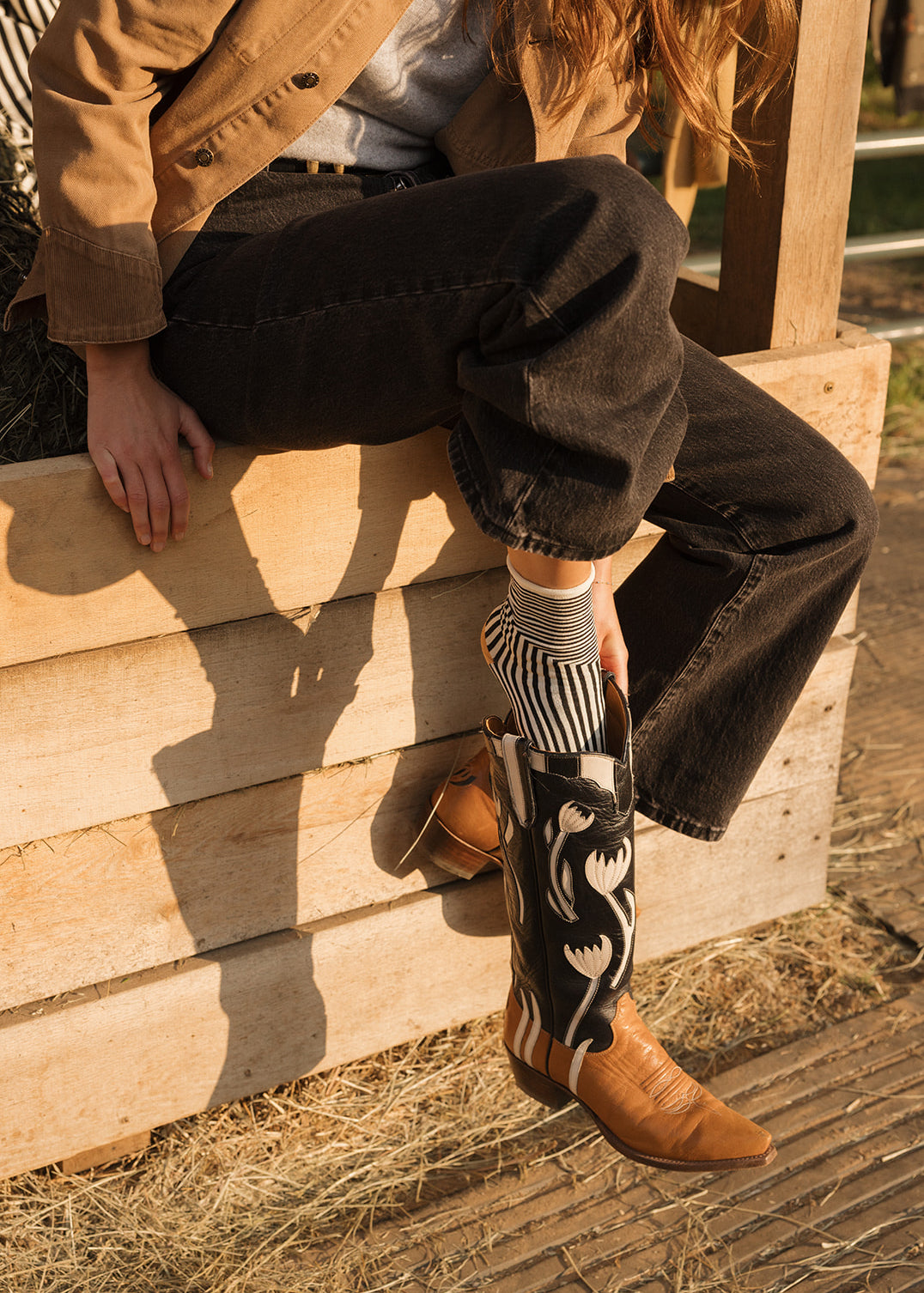 Person wearing cowboy boots and a tan jacket sitting on a wooden structure.