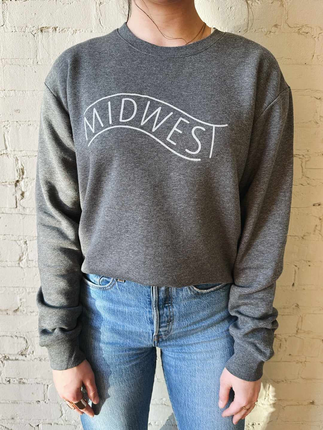 Brunette female model wearing jeans and a grey crewneck sweatshirt with Midwest silkscreened on it.