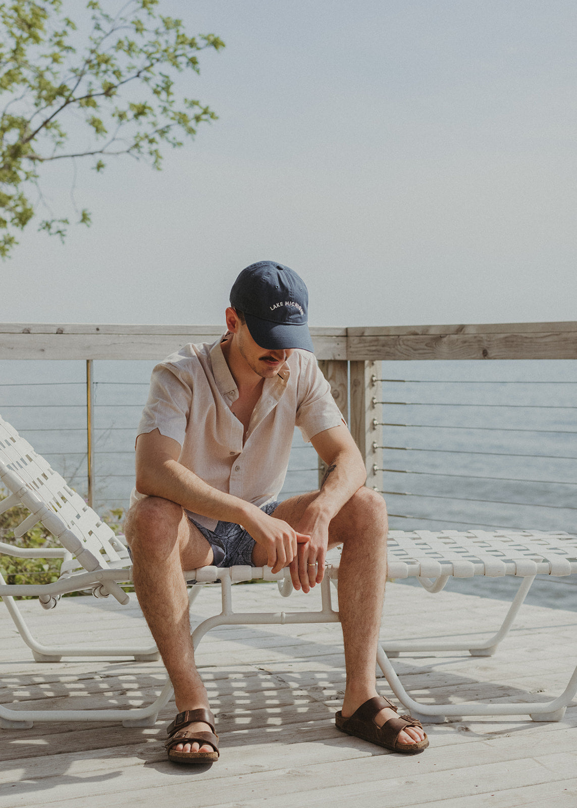 Male model sitting on a beach chair wearing sandals, shorts, a button up shirt and embroidered Lake Michigan dad hat.