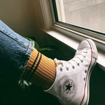 A person wearing gold colored socks with Converse hi-tops and jeans with their toe on a window sil.