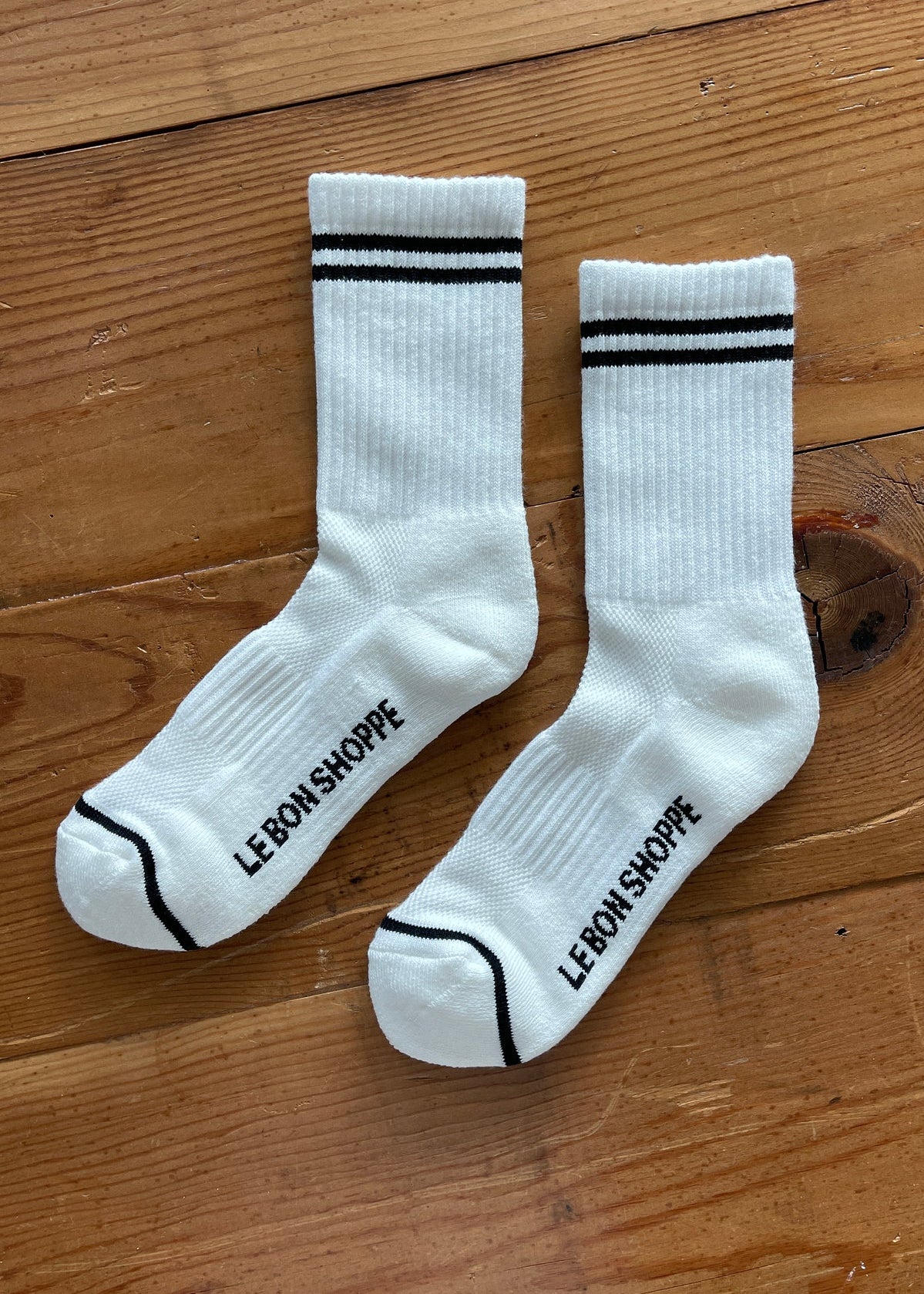 White socks with black stripes on the cuff and Le Bon Shoppe branding on wood surface.