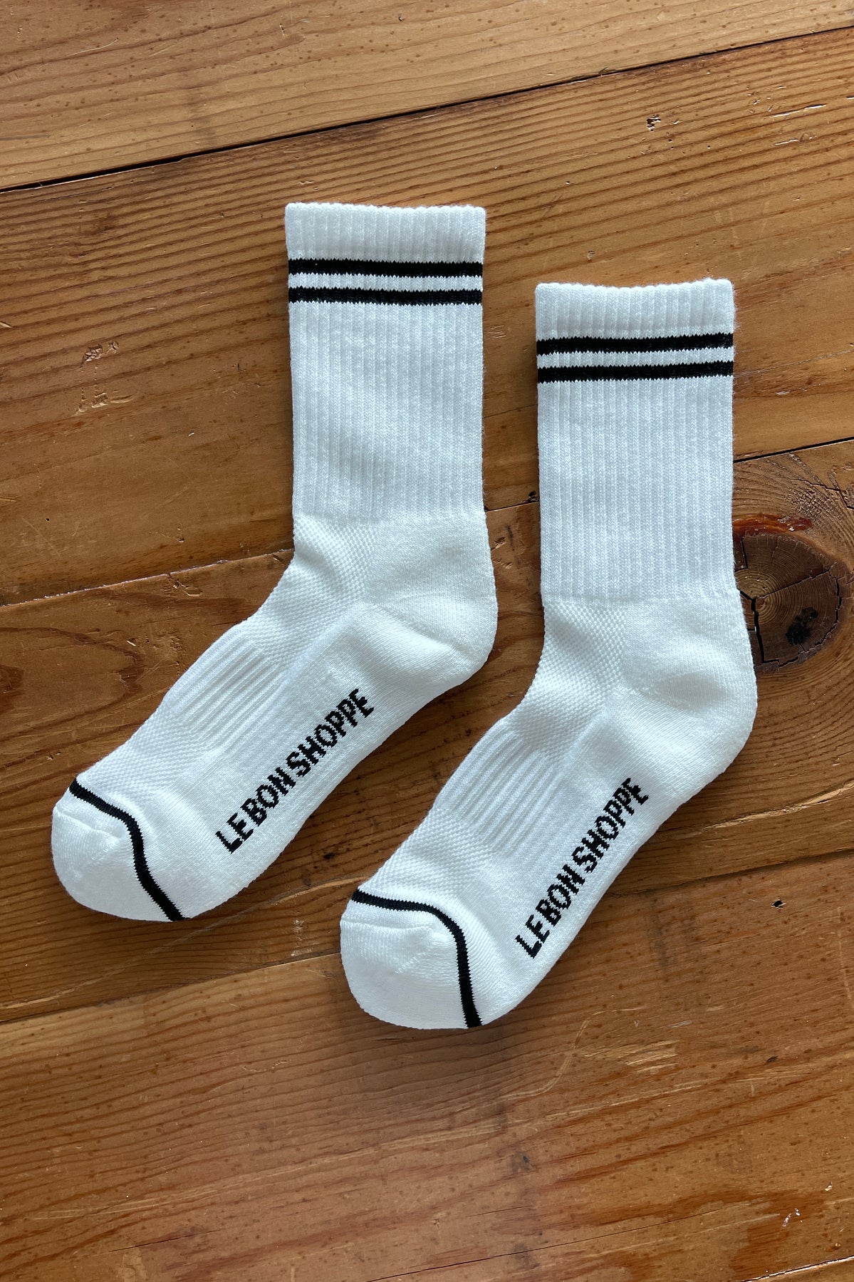 White socks with black stripes on the cuff and Le Bon Shoppe branding on wood surface.