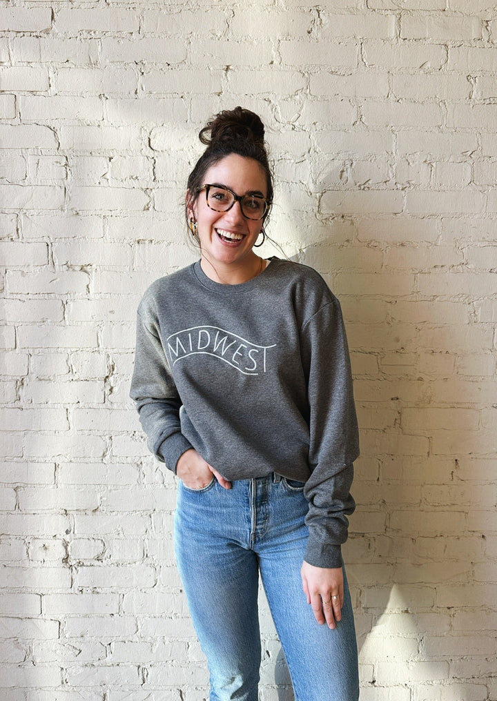 Brunette female model wearing jeans and a grey crewneck sweatshirt with Midwest silkscreened on it.