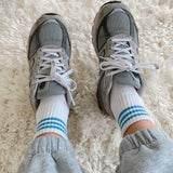 A pair of ivory ankle crew socks with a triple contrast stripe at the opening, worn with grey sneakers.