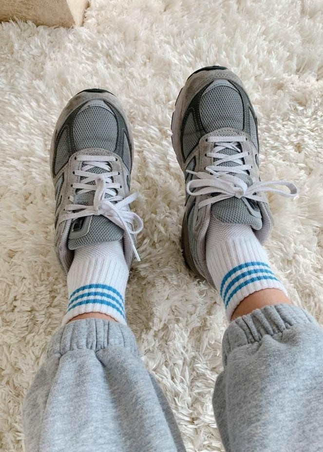 A pair of ivory ankle crew socks with a triple contrast stripe at the opening, worn with grey sneakers.