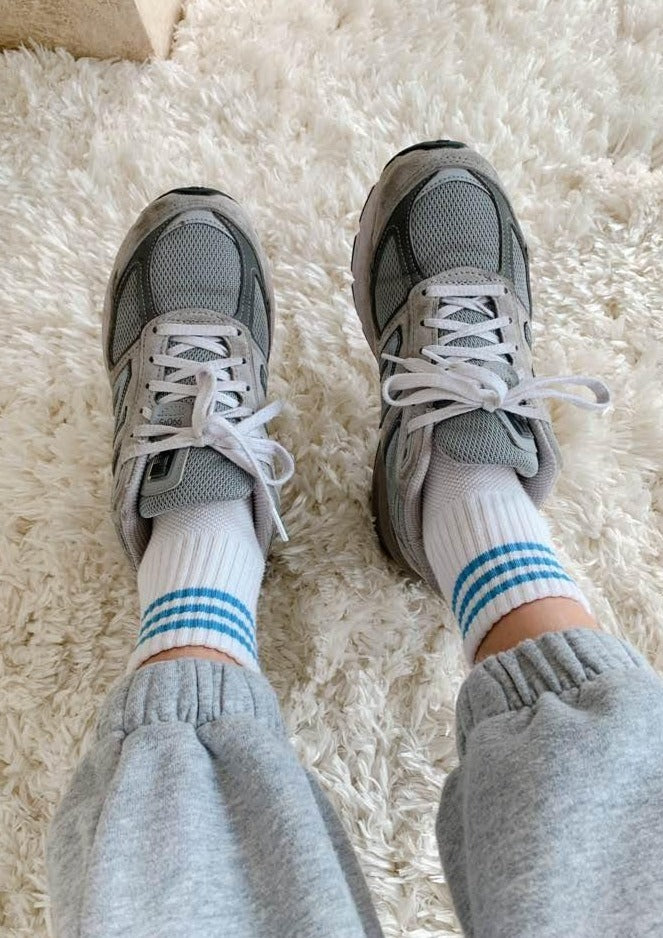 A pair of ivory ankle crew socks with a triple contrast stripe at the opening, worn with grey sneakers.