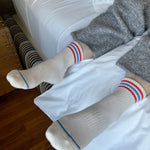 A pair of socks with a white base and red and blue stripes at the cuff, worn on an individual lying on a bed.