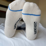 Bottom of person's feet wearing white socks with blue stripe and Le Bon Shoppe branding on white sheets