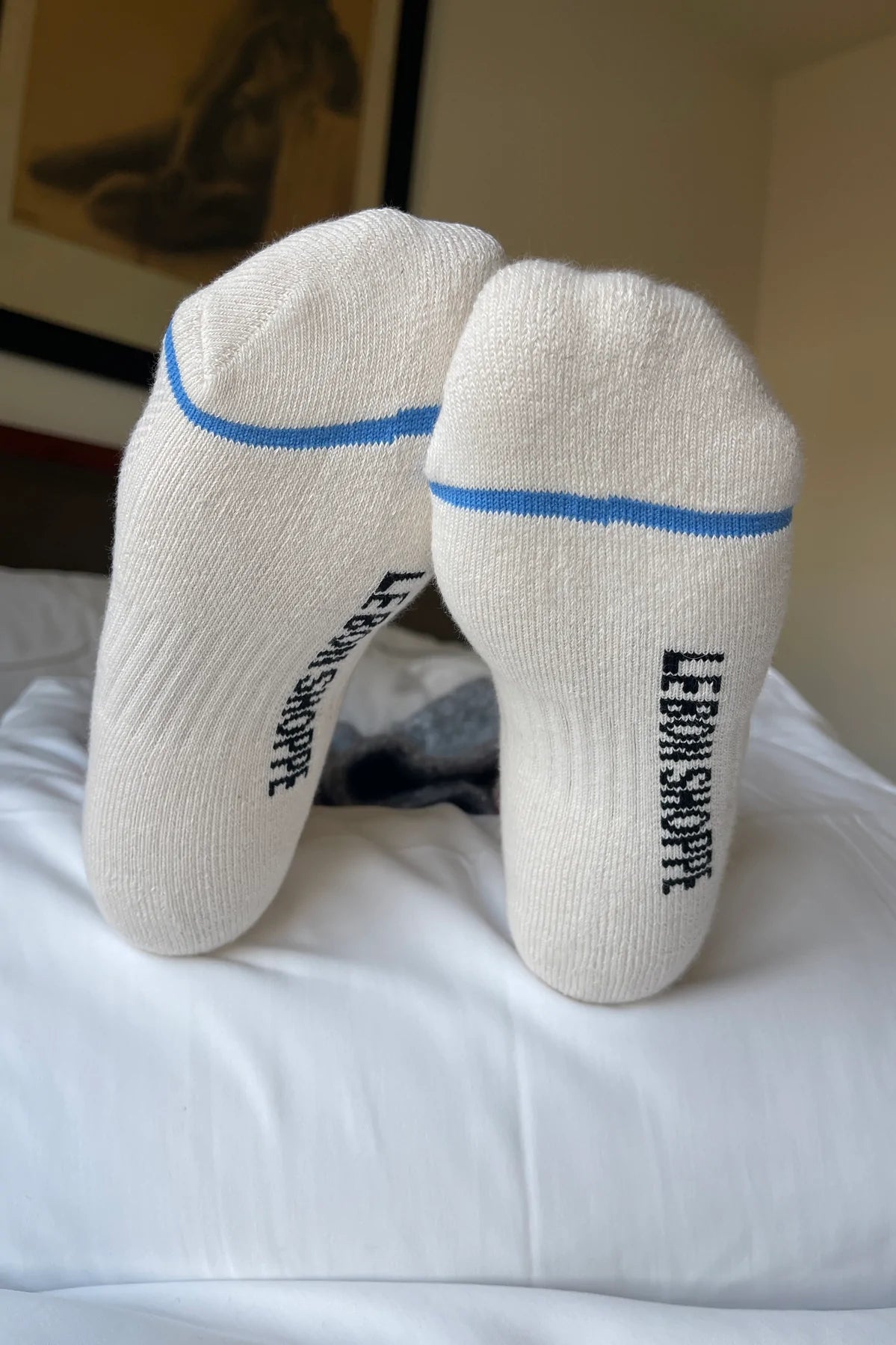 Bottom of person's feet wearing white socks with blue stripe and Le Bon Shoppe branding on white sheets