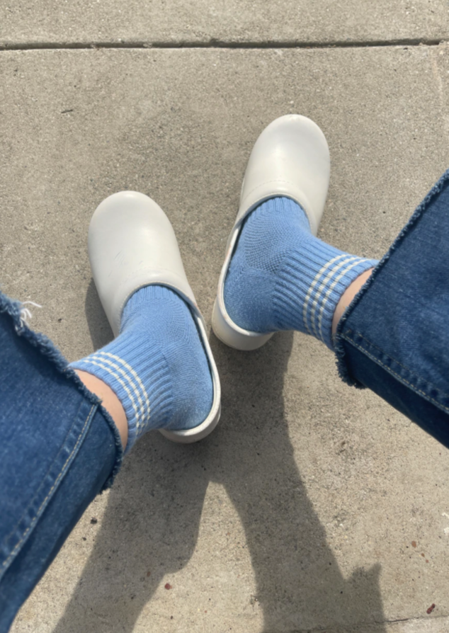 A pair of blue ankle crew socks with a triple contrast stripe at the opening, worn with denim jeans and white clogs.